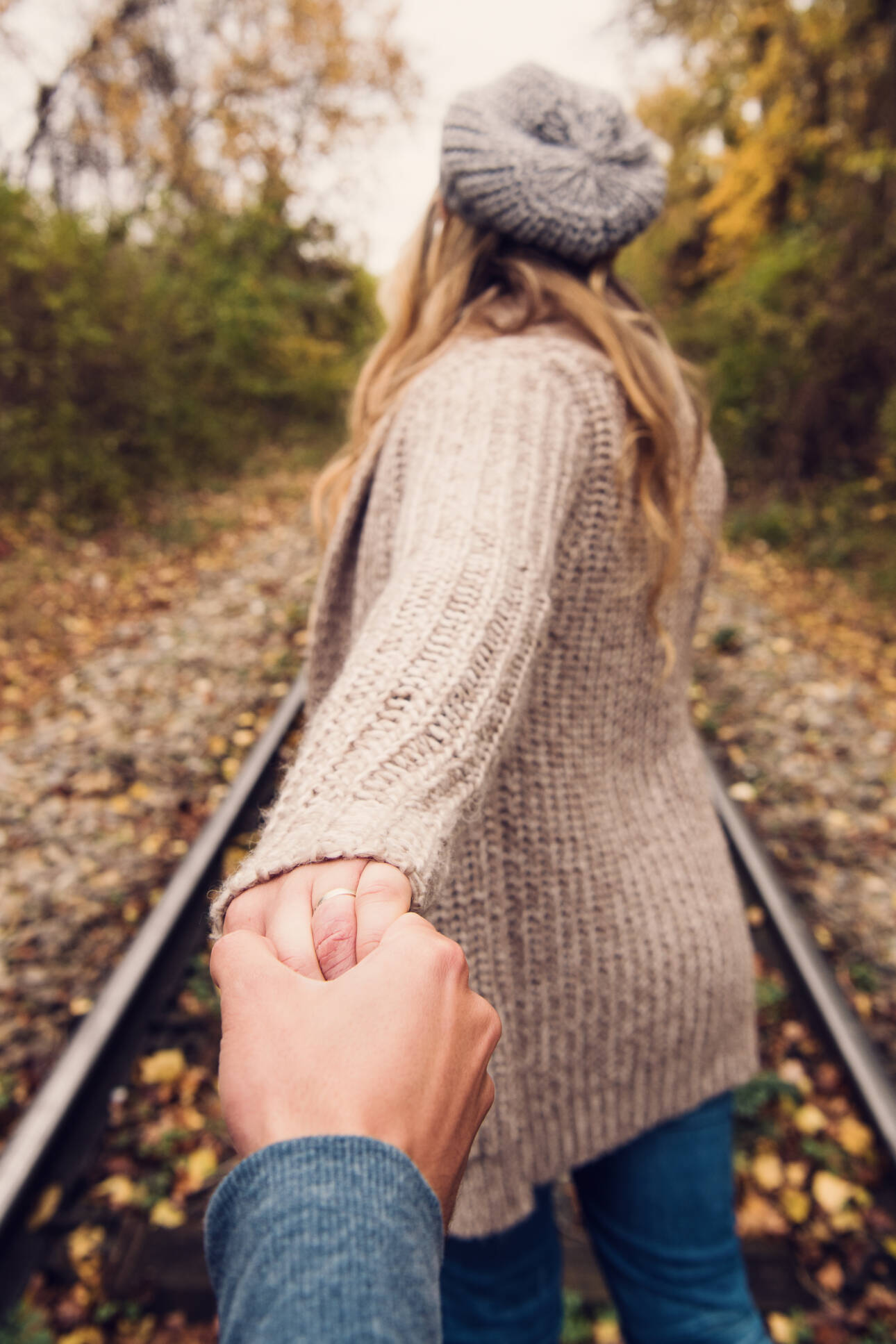 Frau führt ihren Partner an der Hand über herbstliche Bahngleise, Blick aus seiner Perspektive