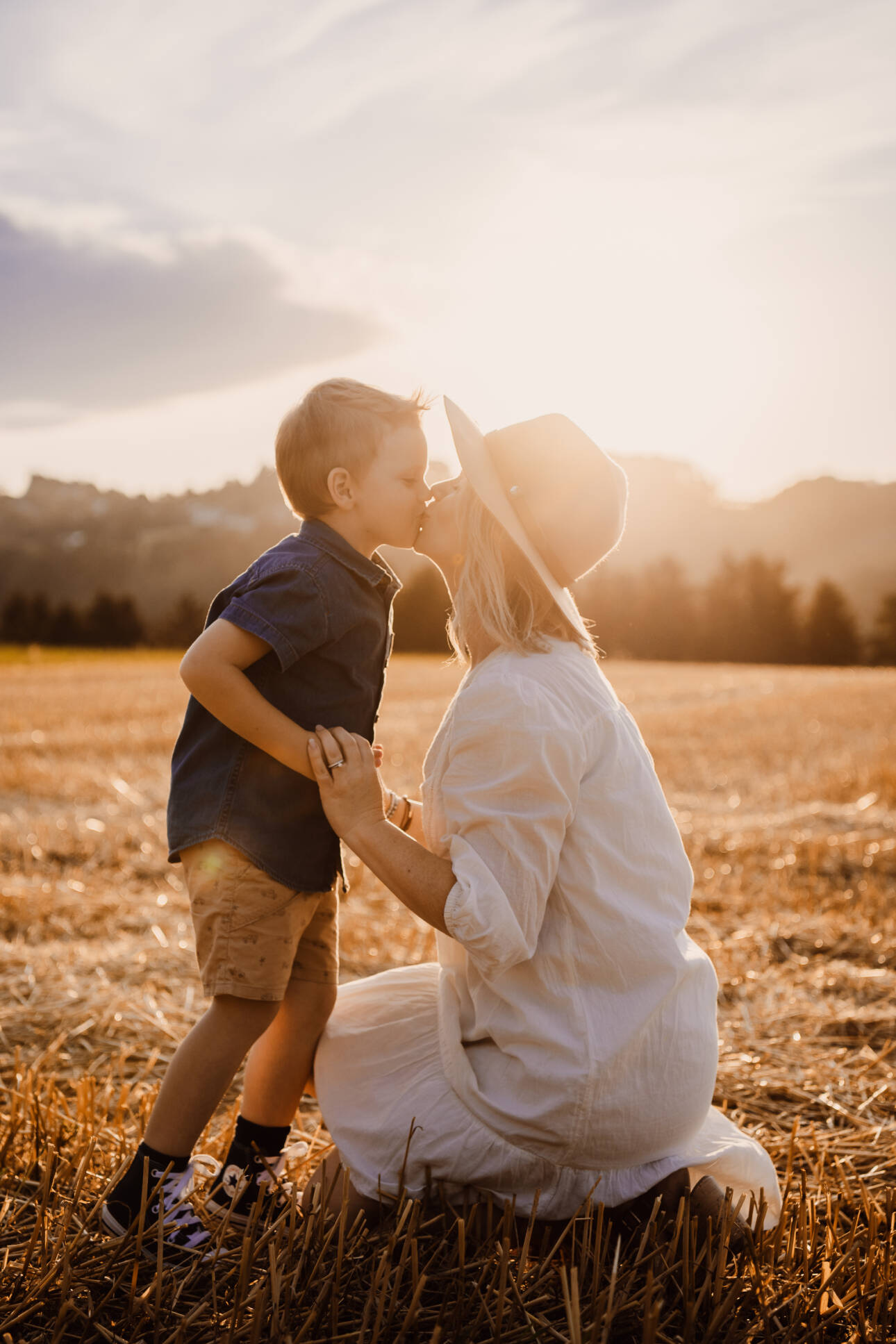 Kniende Mutter und kleiner Sohn küssen sich im Stoppelfeld bei Sonnenuntergang