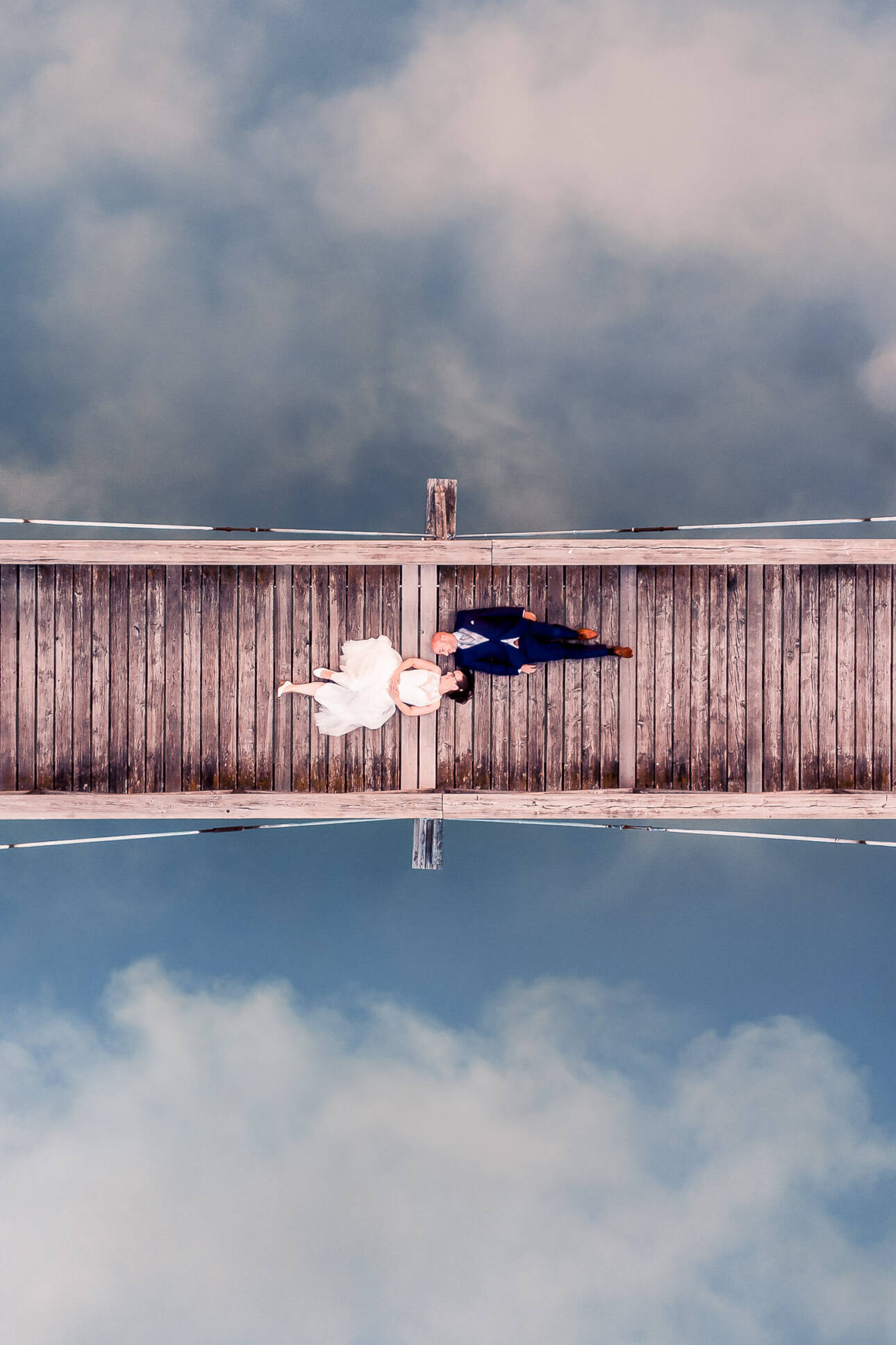 Vogelperspektive auf Brautpaar Hand in Hand auf Holzsteg, Wolken spiegeln sich im Wasser
