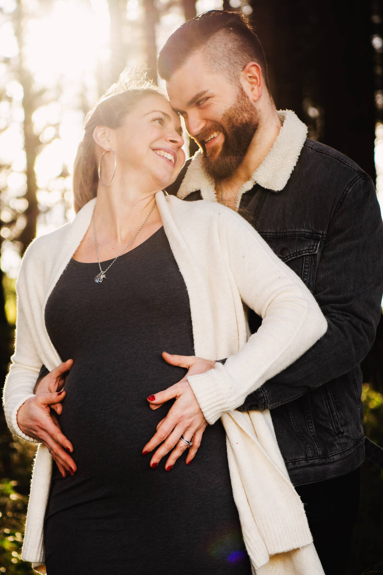 Werdende Eltern halten lachend den Babybauch beim Babybauch-Shooting im sonnendurchfluteten Wald