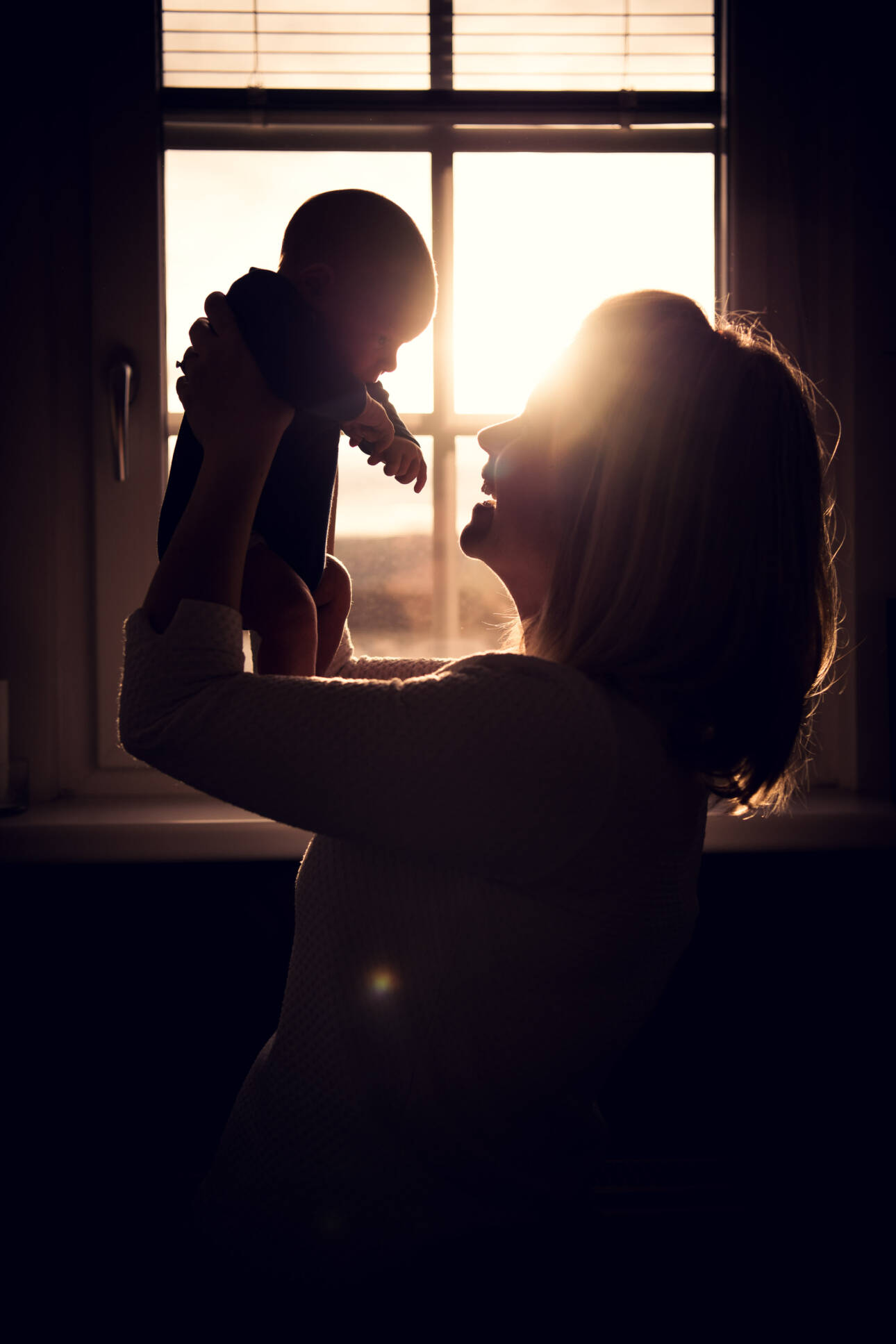 Mutter hält Baby vor sonnendurchflutetem Fenster, warme Silhouette im Gegenlicht